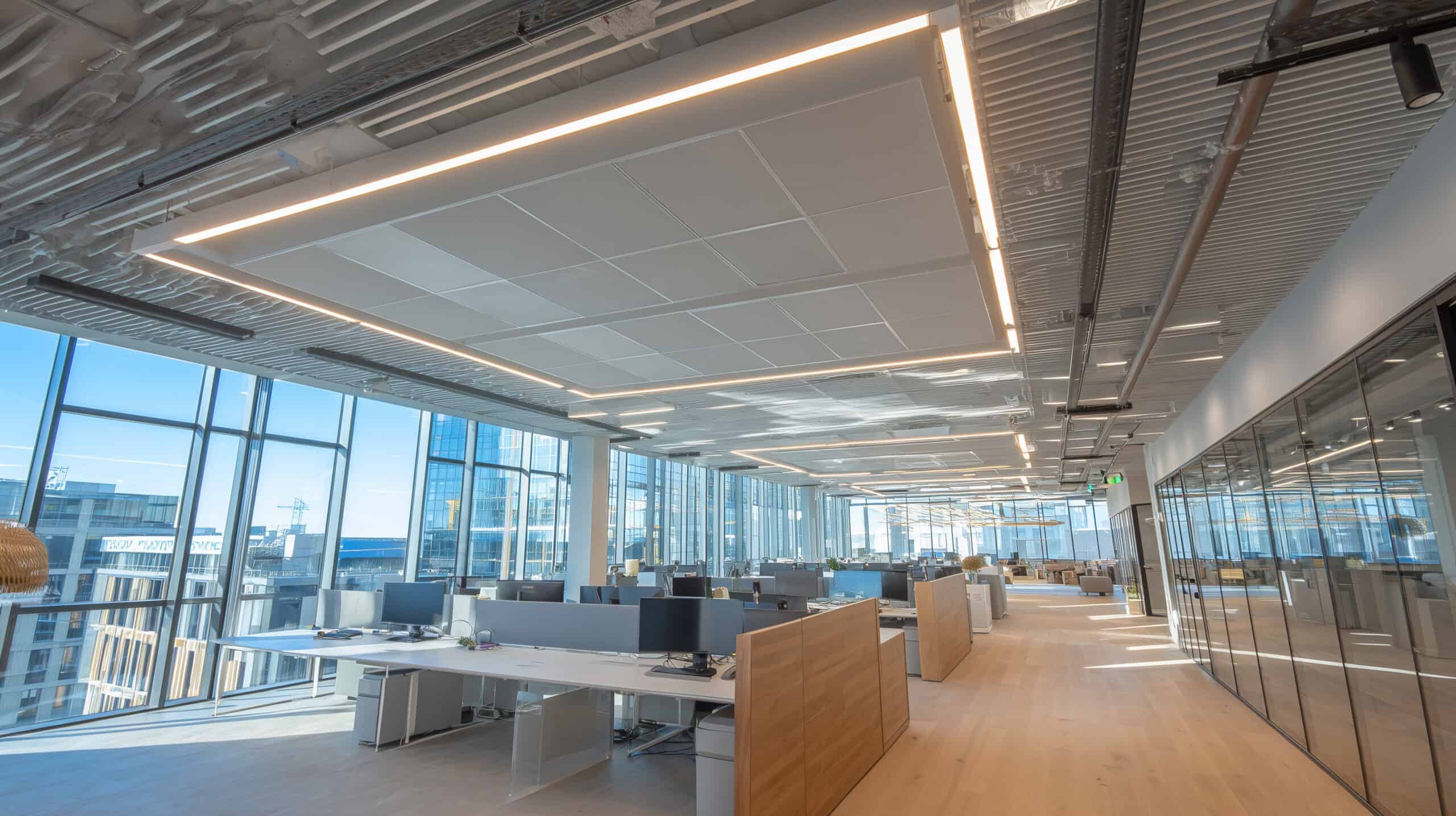 Un bureau moderne à aire ouverte avec de grandes fenêtres, des bureaux avec ordinateurs, des murs en verre, des planchers en bois clair et des plafonds industriels apparents - bien éclairés grâce à un récent relamping LED rénovation éclairage adapté aux bureaux commerciaux.