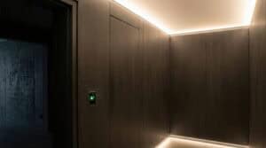 Un couloir minimaliste, faiblement éclairé, avec des murs sombres et un panneau de sécurité intégré doté d'un scanner biométrique vert. Des bandes LED douces éclairent le plafond et le sol, parfaites pour rénover l'éclairage dans les bureaux et commerces et créer une atmosphère élégante et moderne.